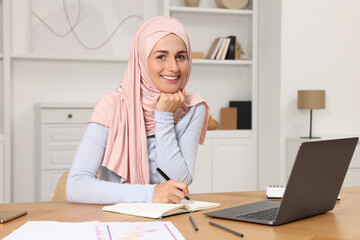 Wall Mural - Muslim woman writing notes near laptop at wooden table in room