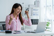 © crizzystudio - Asian businesswoman raising her hand with a happy expression and looking at her laptop.The businesswoman was delighted to receive an email informing her of her annual bonus. concept of success