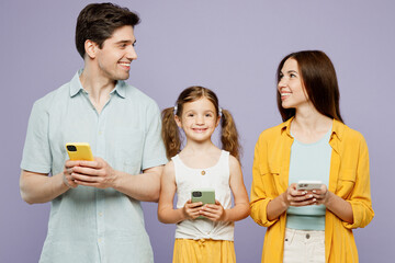 Wall Mural - Young parents mom dad with child kid daughter girl 6 years old wears blue yellow casual clothes hold use mobile cell phone look to each other isolated on plain purple background. Family day concept.