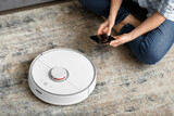 Fototapeta Koty - The girl sits on the floor and controls the robot vacuum cleaner using a smartphone, smart home.