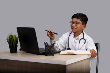 Indian Asian kid boy aged 7 to 8, wearing a doctor apron with stethoscope. with laptop writing prescription. He had a dream to future study as Doctor. He smiled happily, Concept little Doctor