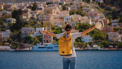 Wall Mural - Nice asian Happy Female with backpack Enjoying her holidays on Symi Islands. View of port Symi or Simi, is tiny island of Dodecanese, Greece, calm atmosphere and fabulous architecture.