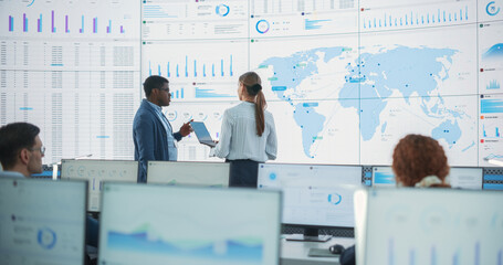 Fototapete - Black Male Analyst Talks To Caucasian Female Manager In Front Of Big Digital Screen With World Map In Monitoring Office. Diverse Employees Working Behind Desktop Computers In Logistics Company.