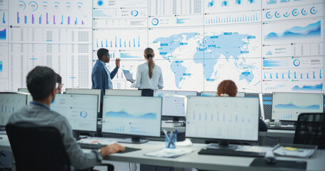 Fototapete - Back View Of Black Male Analyst Talking To Caucasian Female Manager In Front Of Big Digital Screen With World Map In Monitoring Office. Diverse Employees Working Behind Computers In Logistics Company.