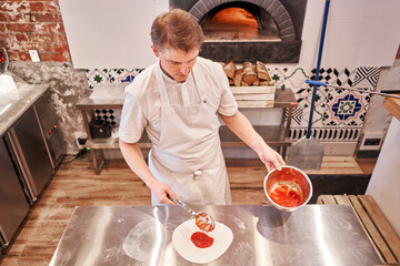 Wall Mural - Cooking pizza. the workpiece poured tomato sauce. Closeup hand of chef baker in uniform apron cook at kitchen