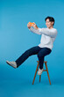 © makistock - Full body of young man sit a stool playing game cellphone isolated over blue background