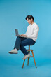 © makistock - Young Asian man sitting and using laptop on bluebackground