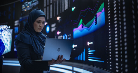 Muslim Female Data Center IT Engineer Standing in a Room with an AI Neural Network Settings on a Digital Screen. Cloud Computing Expert Uses Laptop for Servicing the System in Cyber Security Facility