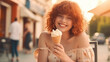 Red-haired curly happy girl eating an ice cream cone against the backdrop of a sunset street.