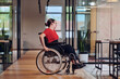 © .shock - A modern young businesswoman in a wheelchair is surrounded by an inclusive workspace with glass-walled offices, embodying determination and innovation in the business world