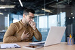 © Tetiana - Asthma attack in the workplace. A young man works in the office at a table with a laptop. He coughs, covers his mouth with his hand, feels weak, sick with a virus