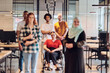 © .shock - A diverse group of young business people congregates within a modern startup's glass-enclosed office, featuring inclusivity with a person in a wheelchair, an African American young man , and a hijab