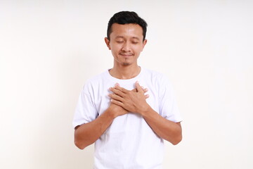 Wall Mural - Relieved asian funny man standing while holding his chest. Isolated on white background