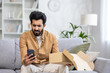 Upset and annoyed man received a parcel, Latin American dissatisfied with the delivery service sits on the sofa in the living room, writes a negative review on the phone.