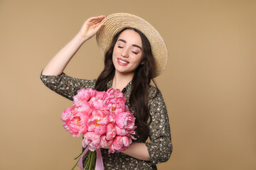 Poster - Beautiful young woman in straw hat with bouquet of pink peonies against light brown background