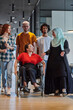 © .shock - A diverse group of young business people walking a corridor in the glass-enclosed office of a modern startup, including a person in a wheelchair and a woman wearing a hijab, showing a dynamic mix of