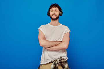 positive man in wireless headphones listening music on blue background, closed eyes, summer fashion