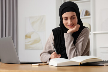 Wall Mural - Muslim woman writing notes near laptop at wooden table in room