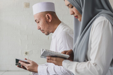 Wall Mural - Asian muslim couple reciting Quran  in mosque during Ramadan. Couple reading holy book of Al Quran together. Selective focus.