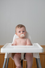 Baby With White Pacifier Sits In White Highchair.