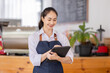 © David - Portrait of asian smiling fun young woman barista barman employee wear brown apron and standing with tablet work in coffee shop. portrait SME Small business entrepreneurs startup concept
