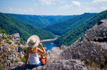 Wall Mural - Woman traveler with hat looking at beautiful mountain,forest and river view- wanderlust travel concept