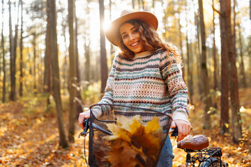 Smiling young woman in a hat and a stylish sweater with a bicycle walks and enjoys the autumn weather in the forest, among the yellow leaves at sunset.