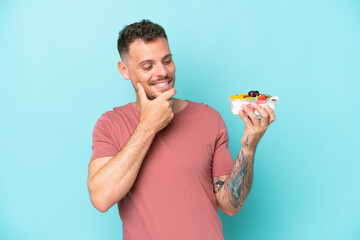 Wall Mural - Young caucasian man holding a bowl of fruit isolated on blue background looking to the side and smiling