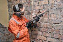 Construction Worker Using Hammer Drill With Chisel To Remove Old Cement From Brick Wall Indoors, Wearing Gloves, Mask, Ear Defenders And Orange Coveralls.