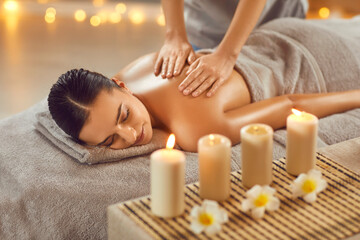 Wall Mural - Relaxed young woman enjoys body massage at spa. Pretty lady getting professional back massage while lying on bed in salon with flowers and aroma candles at luxury resort during blissful summer holiday