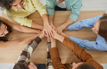 Wall Mural - Top view close up photo of a people hands putting in a stack on wooden table. Young friends or a group of students gathered together and putting their arms in a circle. Unity and teamwork concept.