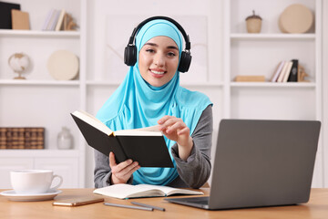 Wall Mural - Muslim woman in headphones studying near laptop at wooden table in room