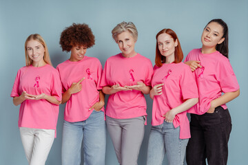 Group of smiling multiracial women showing pink ribbon looking at camera isolated on blue background. Health care, support, prevention. Breast cancer awareness month concept