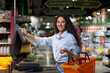 © Liubomir - Portrait of satisfied female shopper in supermarket, Hispanic woman weighing fruit in store, smiling and looking at camera using self-service scales.