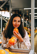 © dvulikaia - Young beautiful dark-haired woman holds oranges in market. Seller of organic fruits at local market. Local business and farm fruits concept.