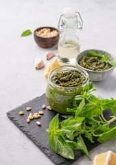 Wall Mural - Traditional Italian pesto in a jar with green basil, pine nuts, cheese and olive oil on a slate board on a light background. A classic sauce for spaghetti or bruschetta.