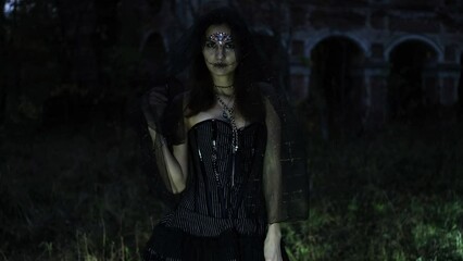 Wall Mural - A mysterious Gothic woman in a black dress with a corset, her head is covered with a black veil. Model posing in front of an old abandoned building. Autumn forest. Halloween concept. Horizontal video