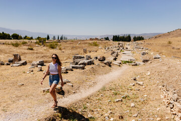 A young woman in a striped top and blue denim shorts walks along a picturesque road between the ruins of an ancient city. Journey to the old city. Hierapolis, Pamukkale, Türkiye - July 29, 2023