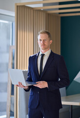 Wall Mural - Confident young adult professional business man company leader, male ceo executive manager wearing formal suit and tie looking at camera holding laptop standing in office. Vertical portrait.