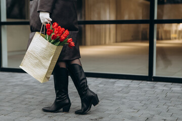 Wall Mural - Cropped image woman walking near glass office building in moody cloudy windy weather with bouquet of red tulips flowers. International Women`s Day 8th March concept
