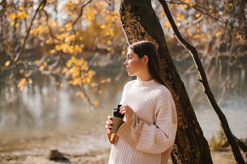 Young brunette woman in a beige sweater is holding an eco glass with coffee in the forest near river in autumn. 