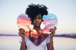 © OpticalDesign - Black woman proud on a sky background, holding an heart shape mirror, self esteem, love and care, beauty and empowerment concept