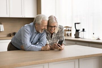 Wall Mural - Happy adorable senior couple watching family pictures, online media content on mobile phone, using smartphone at kitchen table, laughing, feeling joy, talking on video call together