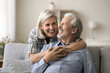 © fizkes - Cheerful caring pretty mature wife hugging happy elder husband from behind with love, tenderness, smiling, laughing. Senior retired couple home casual portrait