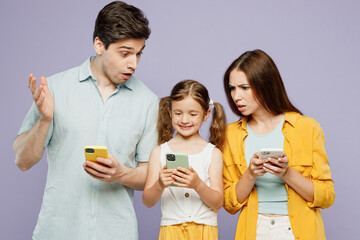 Wall Mural - Young shocked puzzled parents mom dad with child kid girl 6 year old wear blue yellow casual clothes hold use look at daughter mobile cell phone isolated on plain purple background Family day concept.