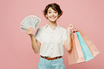 Young smiling woman wear casual clothes hold shopping paper package bags fan of cash money in dollar banknotes isolated on plain pastel pink color background studio. Black Friday sale buy day concept.