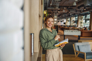 Pretty woman entrepreneur making notes in note pad while standing on cozy coworking background