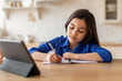 © Prostock-studio - Girl studying at digital tablet writing in notebook at home