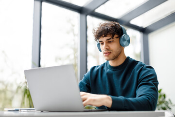 Young european male designer listening music on headphones while working on laptop at a desk in modern glass office
