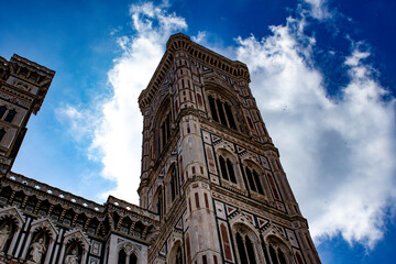 Looking skyward at Giotto's Bell Tower, adjacent to Florence Cathedral, its verticality is emphasized by the cloud-streaked blue sky, showcasing its ornate Gothic architecture.